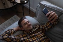 Vue grand angle de l'homme caucasien utilisant le téléphone mobile tout en se penchant sur le canapé à la maison — Photo de stock