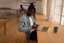 Side view of African-American man using mobile phone with laptop on table at home — Stock Photo