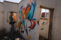 Vue latérale du jeune artiste graffiti caucasien peinture par pulvérisation sur une pièce murale altérée à l'allée — Photo de stock