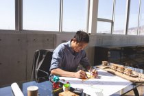 Вид спереди на красивого азиатского архитектора-мужчину, работающего на голубом принте за рабочим столом в современном офисе. Слева от него архитектурная модель . — стоковое фото