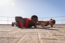 Vista lateral do jovem casal fazendo exercício no pavimento perto da praia do calçadão no dia ensolarado — Fotografia de Stock