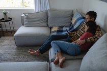 Seitenansicht eines jungen multiethnischen Paares, das sein Handy benutzt, während es sich zu Hause auf dem Sofa lehnt — Stockfoto