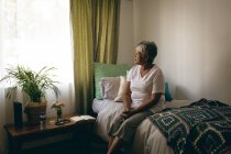 Side view of sad senior mixed race woman sitting on bed at retirement home — Stock Photo