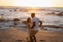 Vista laterale di felice coppia multietnica godendo e giocando sulla spiaggia al tramonto — Foto stock