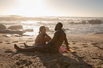 Vista lateral de la joven pareja multiétnica interactuando entre sí en la playa en un día soleado - foto de stock
