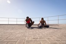 Vue de face du jeune couple multi-ethnique faisant de l'exercice d'étirement sur la chaussée près de la plage de promenade par une journée ensoleillée — Photo de stock