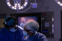 Вид спереду хірургів, що розмовляють один з одним під час операції в операційній кімнаті в лікарні на цифровому екрані на задньому плані — стокове фото