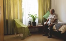 Vue latérale du patient homme caucasien âgé triste regardant par la fenêtre tout en étant assis seul sur le lit de la maison de soins infirmiers — Photo de stock