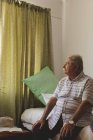 Vista laterale dell'uomo caucasico anziano che guarda fuori dalla finestra mentre è seduto da solo sul letto della casa di cura — Foto stock