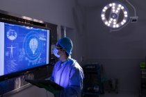 Vue latérale du chirurgien masculin regardant l'écran numérique bleu dans la salle d'opération à l'hôpital — Photo de stock