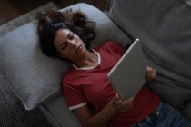 Hochwinkel-Ansicht der schönen Mischlingsfrau mit digitalem Tablet, während sie sich zu Hause auf dem Sofa lehnt — Stockfoto