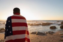 Rückansicht eines afrikanisch-amerikanischen Mannes, der bei Sonnenuntergang am Strand steht und die amerikanische Flagge eingehüllt hat — Stockfoto