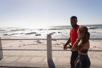 Вид сбоку на молодую многонациональную пару, взаимодействующую друг с другом во время прогулки по тротуару возле пляжа — стоковое фото