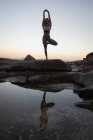 Vista de ángulo bajo de la mujer haciendo yoga sobre roca en la playa al atardecer - foto de stock