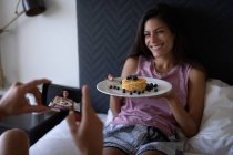 Vista trasera del hombre tomando fotos mientras la mujer de raza mixta sostiene el plato de desayuno en la cama en casa - foto de stock