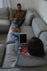 Счастливая многонациональная пара с помощью цифрового планшета и мобильного телефона на диване дома — стоковое фото