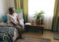 Vista lateral de la reflexiva mujer de raza mixta senior sentada en la cama en el dormitorio de la casa de retiro. Mirando hacia la ventana . - foto de stock