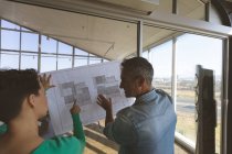 Visão traseira de arquitetos caucasianos segurando o plano contra a janela e discutindo sobre isso no escritório — Fotografia de Stock