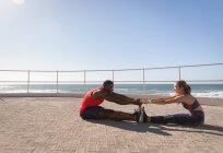Vista laterale della giovane coppia multietnica che fa esercizio di stretching sul marciapiede vicino alla spiaggia sul lungomare in una giornata di sole — Foto stock