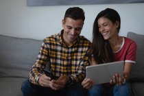 Vista frontal de la feliz pareja multiétnica utilizando el teléfono móvil y la tableta digital en el sofá en casa - foto de stock