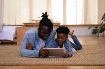 Вид спереди счастливых отца и сына афроамериканцев, которые лежат дома на ковре с помощью цифрового планшета — стоковое фото