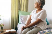 Side view of thoughtful senior mixed race woman sitting on bed and looking at window at retirement home — Stock Photo