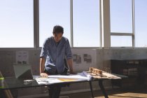 Vista frontal del arquitecto asiático de pie en el escritorio y mirando el plano en una oficina moderna - foto de stock