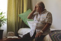 Vista lateral do homem caucasiano sênior olhando fora da janela enquanto sentado sozinho e segurando a cabeça na cama do lar de idosos — Fotografia de Stock