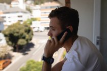Вид сбоку на молодого кавказца, разговаривающего по мобильному телефону, стоя дома на балконе — стоковое фото