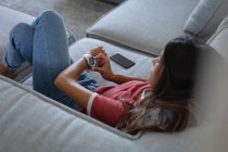 Hochwinkelaufnahme einer Frau mit gemischter Rasse, die Smart Watch benutzt, während sie sich zu Hause auf dem Sofa lehnt — Stockfoto