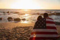 Вид спереду молода багатоетнічна пара загорнула американський прапор, розслабившись на пляжі під час заходу сонця. Вони виглядають горизонтально — стокове фото