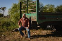 Vista frontal del hombre caucásico mayor esperando mientras está sentado en el remolque en la granja en un día soleado - foto de stock