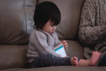 Vue de face d'un petit bébé asiatique mignon jouant avec un téléphone portable assis sur le canapé à côté de sa mère à la maison — Photo de stock