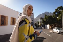 Вид збоку жінки змішаної раси з використанням мобільного телефону під час прогулянки по середині вулиці в сонячний день — стокове фото