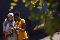 Vista frontal de jóvenes amigas tomando selfie con teléfono móvil en la calle de la ciudad - foto de stock