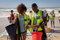 Frontansicht multiethnischer Freiwilliger, die an einem sonnigen Tag über Klemmbrett am Strand diskutieren — Stockfoto