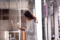 Seitenansicht einer asiatischen Frau, die ihr Mobiltelefon benutzt, während sie in einem Glaskorridor steht — Stockfoto
