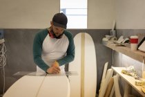 Vorderansicht eines gutaussehenden kaukasischen Mannes mit Ohrenschützern, der mit Lineal und Bleistift ein Surfbrett in der Werkstatt misst — Stockfoto