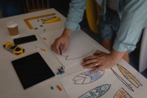 Der mittlere Abschnitt des Menschen zeichnet Surfbrett-Skizze in einem Workshop. Tablet, Kaffee, Lineal, Bleistifte, Buntstifte, Papier und Handy liegen auf dem Tisch. — Stockfoto