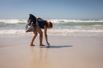 Vista lateral de la joven mujer blanca voluntaria limpiando la playa en un día soleado. Está tocando la arena . - foto de stock