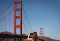 Vue arrière de la femme capturant l'image avec le téléphone portable du pont suspendu — Photo de stock