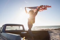 Вид спереди красивой молодой афроамериканки, держащей в руках американский флаг, стоя на машине на пляже в солнечный день — стоковое фото