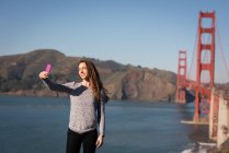Vista frontale della bella donna che prende selfie di fronte al ponte sospeso — Foto stock