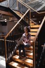 Вид збоку красивою змішаної раси ділової жінки розмова на мобільному телефоні, працюючи на ноутбуці на сходах в сучасному офісі — стокове фото