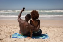 Vista posteriore di felice coppia multietnica scattare selfie con il telefono cellulare sulla spiaggia — Foto stock