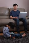 Зовнішній вигляд молодого азіатського батька з використанням ноутбука на дивані, а його син за допомогою цифрової таблетки на дому — стокове фото