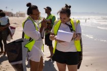 Vista frontal de mujeres voluntarias multiétnicas felices discutiendo sobre el portapapeles mientras el otro voluntario está limpiando en la playa - foto de stock