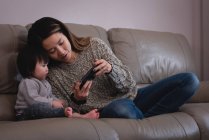 Vista frontale di madre e figlia asiatica utilizzando il telefono cellulare mentre si siede sul divano a casa — Foto stock
