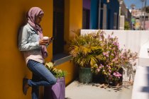 Vista laterale di bella donna di razza mista con hijab utilizzando il telefono cellulare mentre prende il caffè a casa in una giornata di sole — Foto stock