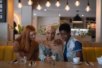 Vista frontal de felices amigas de raza mixta usando teléfono móvil mientras están sentadas en el restaurante - foto de stock